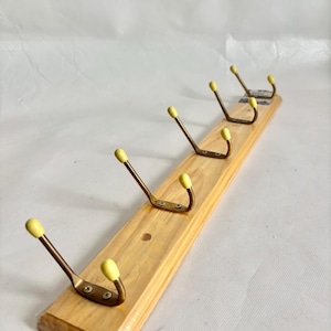 May include: A wooden coat rack featuring five brass hooks with yellow tips, mounted on a light-coloured wooden board. The rack is designed for hanging coats, hats, or other items. The board measures approximately 60 cm in length.