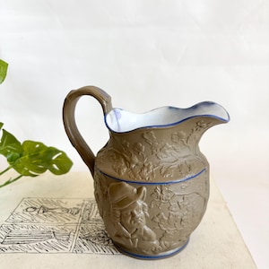 Puede incluir: Una jarra de cerámica marrón con asa y un diseño en relieve detallado. La jarra presenta un hombre con sombrero y motivos florales. El borde es festoneado y ribeteado en azul, y el interior es blanco.