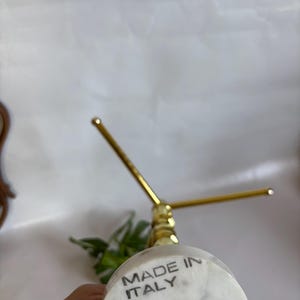 May include: A marble and brass jewelry display stand. The base is white marble with the text "MADE IN ITALY". The stand has a gold-colored, three-pronged top. The stand is designed to hold jewelry.
