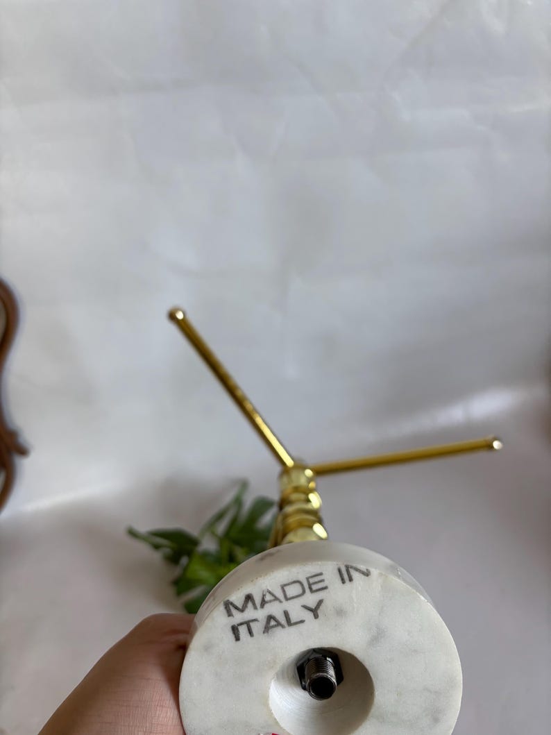 May include: A marble and brass jewelry display stand. The base is white marble with the text "MADE IN ITALY". The stand has a gold-colored, three-pronged top. The stand is designed to hold jewelry.