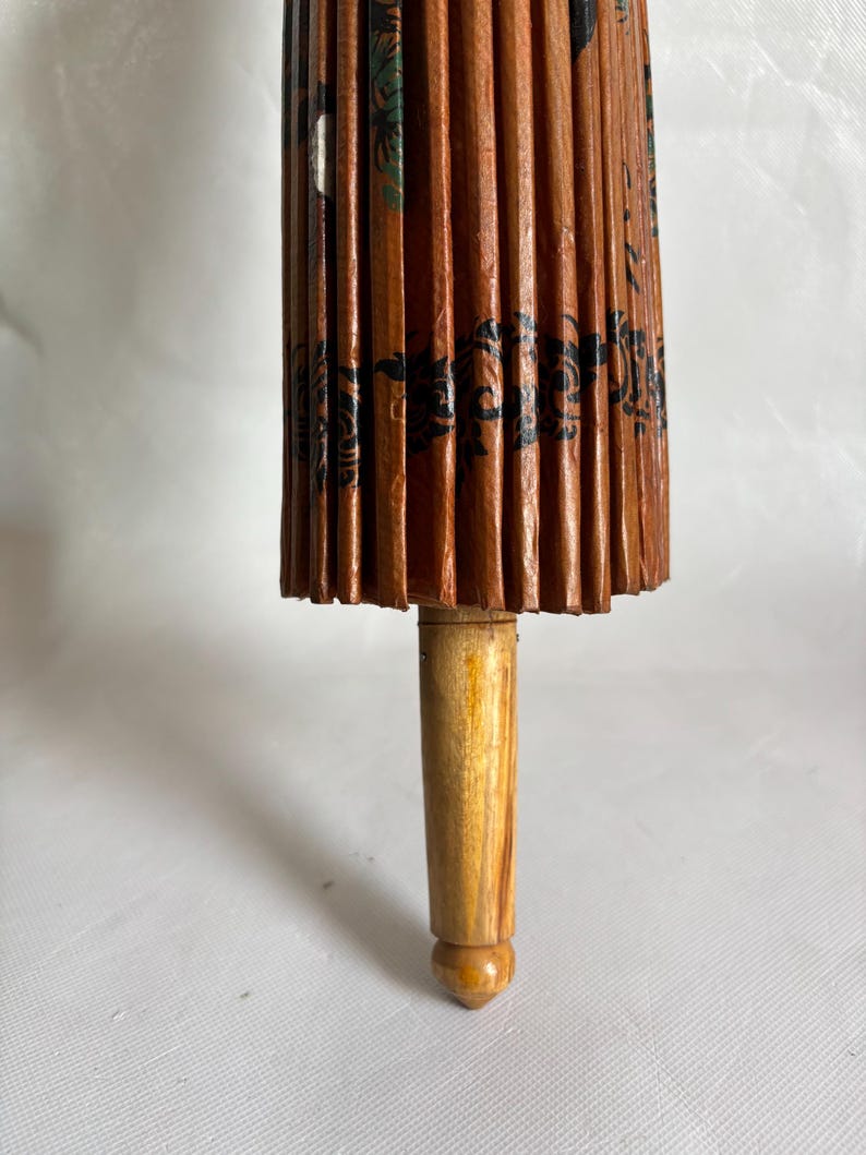 Puede incluir: Primer plano de un parasol japon&eacute;s tradicional. El parasol est&aacute; hecho de papel marr&oacute;n con dise&ntilde;os florales negros y verdes. El mango es de madera clara.