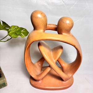 May include: A smooth, light brown terracotta sculpture depicting two abstract figures embracing. The figures have rounded, featureless heads and curved, flowing bodies that form a circular, interconnected shape with a central heart-like void.