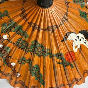 Puede incluir: Parasol de papel naranja con un mango negro y obras de arte decorativas. La obra de arte incluye &aacute;rboles verdes, p&aacute;jaros blancos y un sol rojo. Tambi&eacute;n hay patrones decorativos negros.