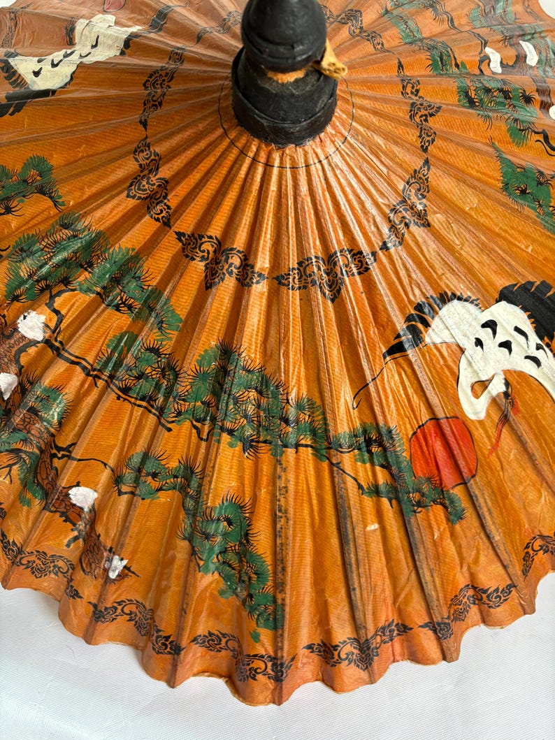 Puede incluir: Parasol de papel naranja con un mango negro y obras de arte decorativas. La obra de arte incluye &aacute;rboles verdes, p&aacute;jaros blancos y un sol rojo. Tambi&eacute;n hay patrones decorativos negros.