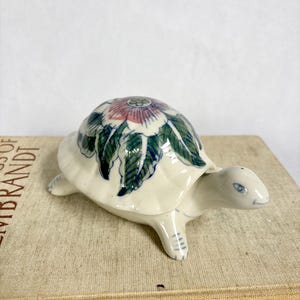 May include: A ceramic turtle figurine with a floral design on its shell. The turtle is white with green, blue, and pink accents. The figurine is resting on a book.