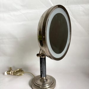 Puede incluir: Un espejo de tocador de estilo vintage con un marco circular iluminado. El espejo tiene un acabado de metal plateado, una columna veteada en negro y gris y una base redonda. Un cable de alimentación se extiende desde la base.