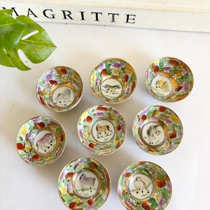 May include: Eight small, round porcelain bowls with a gold rim and floral design. Each bowl features a different horse illustration in the center. The bowls are arranged on a white surface, with a book titled "MAGRITTE" in the background.