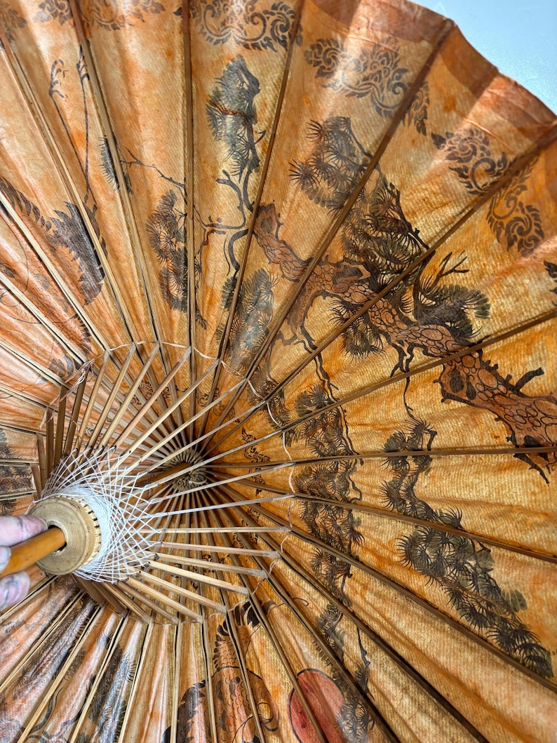 Puede incluir: Parasol japon&eacute;s antiguo, abierto, con un esquema de color naranja c&aacute;lido y marr&oacute;n. El interior presenta una ilustraci&oacute;n detallada de &aacute;rboles y follaje. Las varillas y el mango de madera son visibles. El parasol est&aacute; parcialmente abierto.