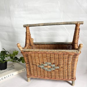May include: Wicker magazine rack with a handle. The basket is woven with natural and light brown wicker, accented with light blue-gray details. The rack has a rectangular shape with a decorative diamond pattern on the front.