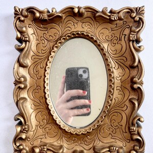 Puede incluir: Espejo ovalado en un marco dorado ornamentado. El marco tiene un dise&ntilde;o detallado de volutas. El espejo refleja una mano que sostiene un tel&eacute;fono. Un art&iacute;culo decorativo.