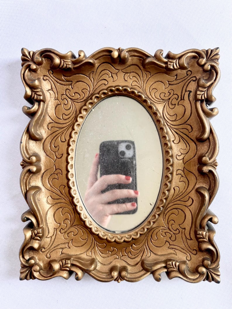 Puede incluir: Espejo ovalado en un marco dorado ornamentado. El marco tiene un dise&ntilde;o detallado de volutas. El espejo refleja una mano que sostiene un tel&eacute;fono. Un art&iacute;culo decorativo.