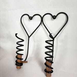 May include: Two black metal wine bottle stoppers with heart-shaped tops and spiral designs. The stoppers have wooden bases and are set against a white background. The design is simple and elegant.