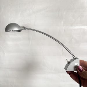 May include: A silver-colored desk lamp with a curved arm and a rounded lamp head. The lamp has a small base with a switch and a black power cord. The lamp is held by a person.
