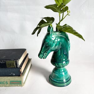 Puede incluir: Un jarrón de cerámica verde con forma de cabeza de caballo y acabado brillante, que contiene tallos verdes y frondosos. El jarrón tiene forma de pieza de ajedrez de caballo. Una pila de libros es visible en el fondo.