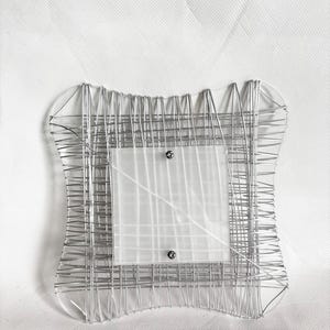 Peut inclure: Un cadre photo décoratif de forme carrée. Le cadre est constitué de fil d'argent, tissé selon un motif en forme de grille. Un insert en verre dépoli est fixé au centre avec deux petites vis. Le cadre a un design artistique unique.