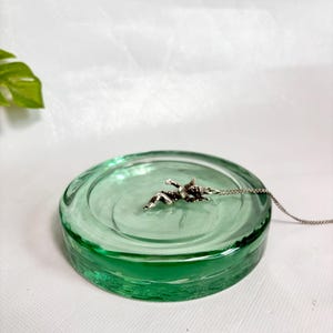 Peut inclure: Un pendentif argenté en forme de petite figurine ailée attaché à une chaîne délicate, reposant à l'intérieur d'un plat circulaire en verre vert clair translucide avec une surface lisse légèrement concave et un bord épais arrondi.