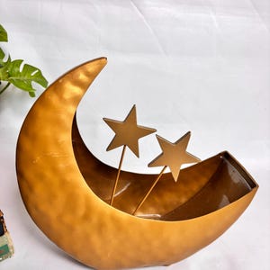 Op de afbeelding: Een halvemaanvormige metalen schaal met een gehamerde gouden afwerking. Twee vijfpuntige sterornamenten op dunne metalen stelen zijn aan de binnenkant van de schaal bevestigd. De schaal heeft een gladde, donkere binnenkant en een gouden buitenkant.