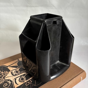 May include: A black plastic desk organizer with multiple compartments of varying sizes. The organizer is sitting on top of a brown book with a decorative design on the cover. The organizer is designed to hold pens, pencils, and other office supplies.