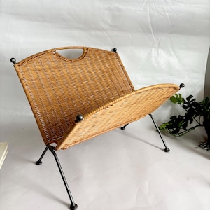 May include: A vintage magazine rack made of woven wicker in a light brown color. The rack has a curved top with a handle and is supported by black metal legs. The legs have rounded feet. The overall design is mid-century modern.