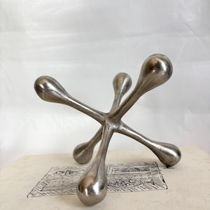 May include: A silver-colored metal sculpture in the shape of a jack, with six teardrop-shaped ends. The sculpture is resting on a book with a black and white illustration.