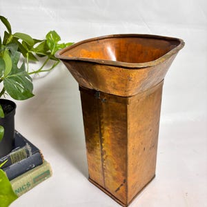May include: A tall, rectangular copper vessel featuring a wide, flared, square-shaped opening at the top. The metal surface displays a weathered, warm orange-brown patina with visible seams along the vertical edges and a smooth, polished interior.