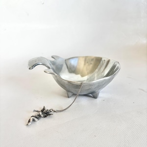May include: A silver-colored tea infuser with a cherub charm attached by a chain. The infuser has a bowl shape with a handle and three small feet. The cherub charm is detailed with wings and a serene expression.