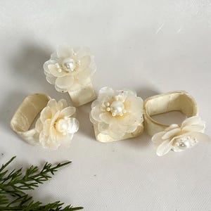 Könnte beinhalten: Vier elfenbeinfarbene Serviettenringe mit floralen Verzierungen. Jeder Ring hat eine Stoffblume mit einem Perlenmittelpunkt. Die Blumen haben mehrere Blütenblattschichten. Die Serviettenringe sind auf einer weißen Oberfläche angeordnet.