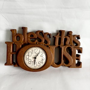 May include: Wooden wall clock with the words "bless this HOUSE" carved into the wood. The clock face is white with black numbers and hands. The clock is a warm brown color.