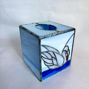 May include: A stained glass tissue box in shades of blue and white, featuring a swan design. The box is square with a circular opening on top. The swan has white feathers, a red beak, and is set against a blue water background.