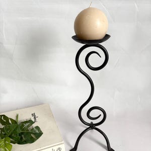 Könnte beinhalten: Ein schwarzer Kerzenhalter aus Schmiedeeisen mit einer runden, cremefarbenen Kerze. Der Kerzenhalter hat ein dekoratives, wirbelndes Design und einen dreibeinigen Sockel. Der Kerzenhalter ist etwa 30 cm hoch.