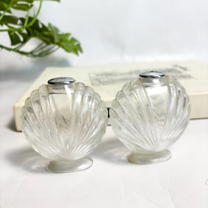 May include: Two clear glass salt and pepper shakers. Each shaker has a shell-like design with a silver metal top and a small, round base. The shakers are on a white surface with a green plant in the background.