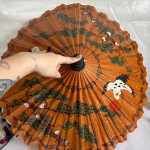 Puede incluir: Un parasol naranja con un mango negro y un dise&ntilde;o pintado detallado. El dise&ntilde;o incluye &aacute;rboles verdes, p&aacute;jaros blancos y acentos rojos. El parasol est&aacute; abierto y abanicado, mostrando la intrincada obra de arte.