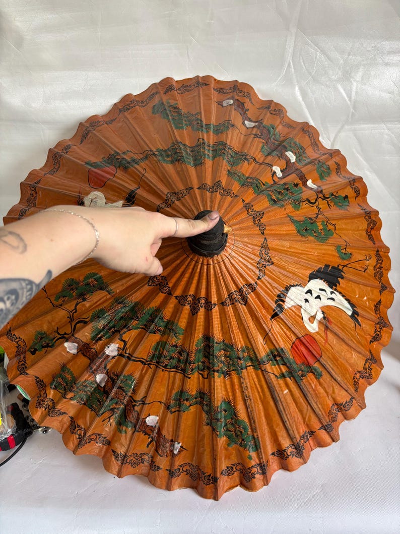 Puede incluir: Un parasol naranja con un mango negro y un dise&ntilde;o pintado detallado. El dise&ntilde;o incluye &aacute;rboles verdes, p&aacute;jaros blancos y acentos rojos. El parasol est&aacute; abierto y abanicado, mostrando la intrincada obra de arte.