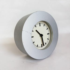 Puede incluir: Un reloj analógico redondo de color plateado con esfera blanca y manecillas e indicadores negros. El reloj tiene un diseño sencillo y moderno sobre un fondo blanco.