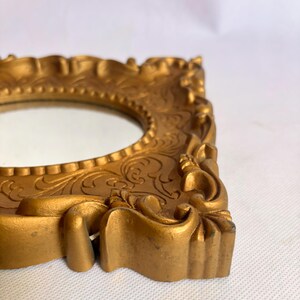 Puede incluir: Un marco de fotos dorado y ornamentado con una abertura ovalada. El marco tiene un dise&ntilde;o en relieve detallado y un acabado ligeramente envejecido. El marco probablemente est&aacute; hecho de resina o un material similar y es adecuado para exhibir una foto peque&ntilde;a o una obra de arte.