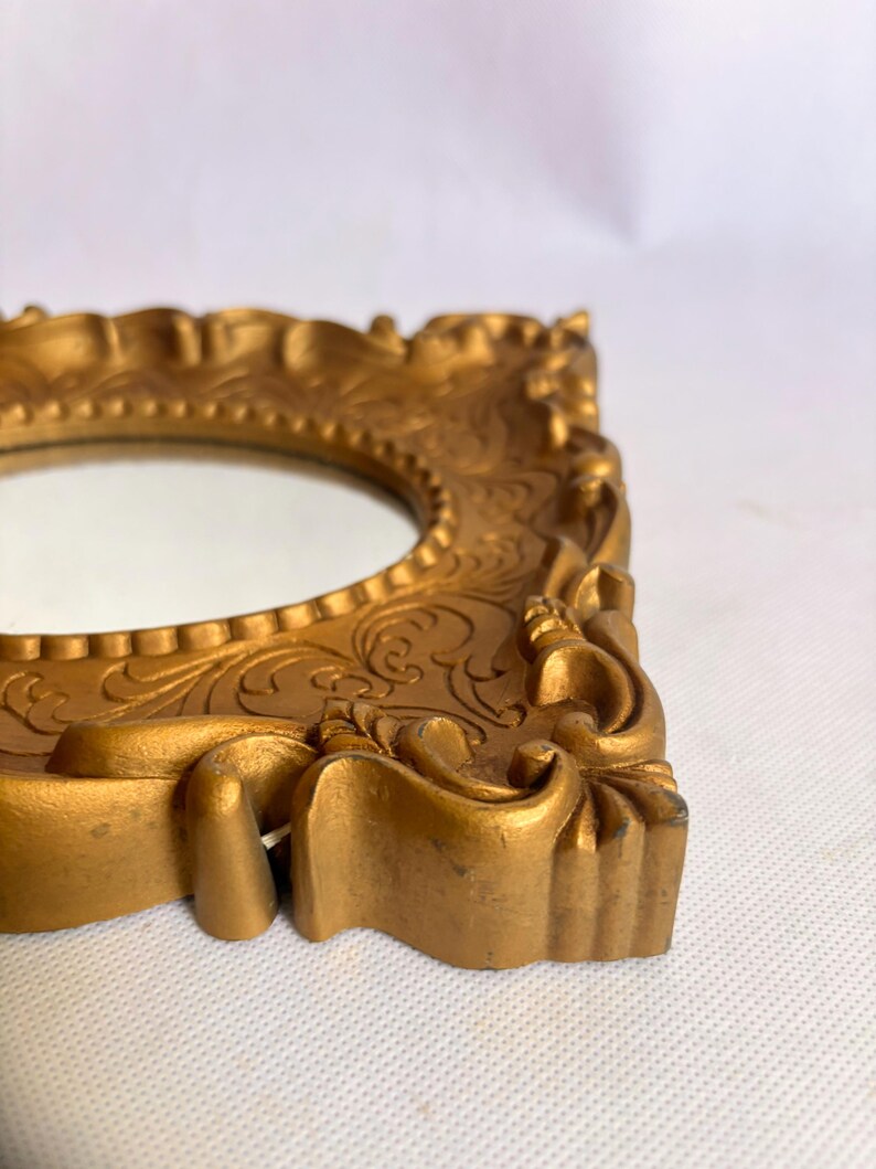Puede incluir: Un marco de fotos dorado y ornamentado con una abertura ovalada. El marco tiene un dise&ntilde;o en relieve detallado y un acabado ligeramente envejecido. El marco probablemente est&aacute; hecho de resina o un material similar y es adecuado para exhibir una foto peque&ntilde;a o una obra de arte.
