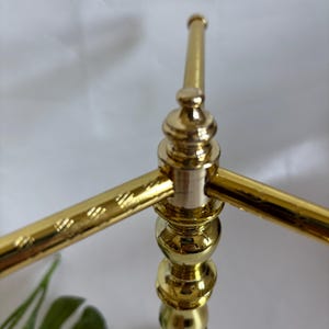 May include: A gold-colored metal decorative stand with a marble base. The stand has a three-arm design with a series of decorative spheres and a textured pattern on the arms. The stand is designed to hold items.