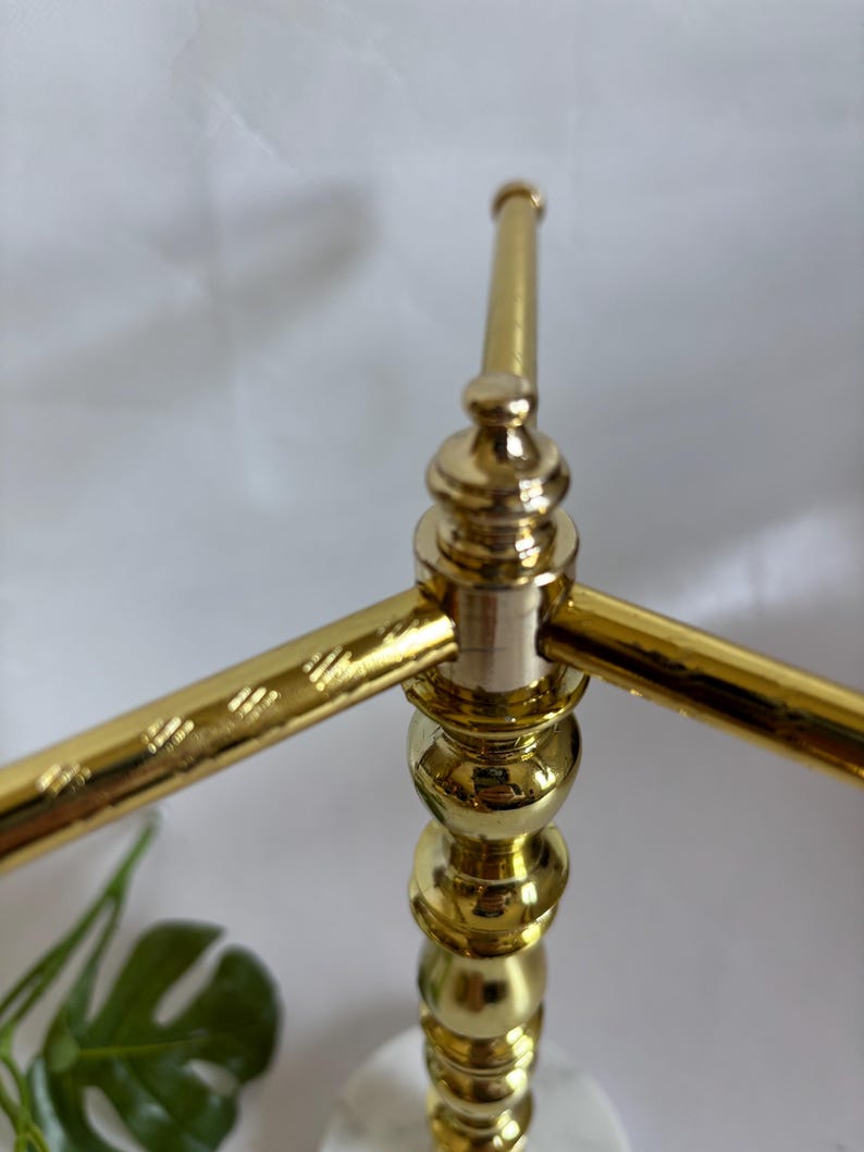 May include: A gold-colored metal decorative stand with a marble base. The stand has a three-arm design with a series of decorative spheres and a textured pattern on the arms. The stand is designed to hold items.