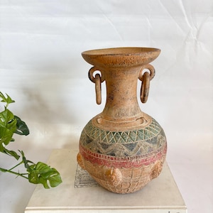 May include: A terracotta-colored vase with a wide rim and two loop handles. The vase features intricate patterns in green, black, and red around the body. The vase is sitting on a book with the title "MAGRITTE".