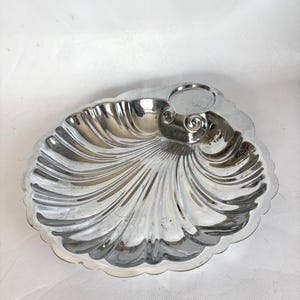 Op de afbeelding: Een zilverkleurige, schelpvormige schaal met een geschulpte rand. De schaal heeft een gedetailleerd, geribbeld ontwerp dat vanuit het midden uitstraalt. Aan de zijkant bevindt zich een kleine, cirkelvormige inkeping. De schaal is waarschijnlijk van metaal.