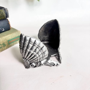 May include: A silver-toned metal trinket box shaped like an open scallop shell. The exterior features a textured, ribbed shell pattern with ornate, swirling decorative details at the base. The interior is hollow and dark, with a hinged opening mechanism.