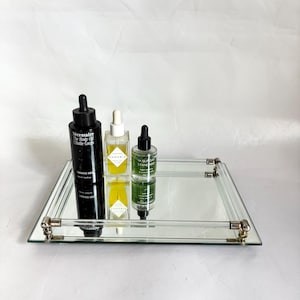 May include: A rectangular mirrored tray with clear glass sides and silver-toned metal accents. Three glass bottles with black droppers and one with a white dropper are arranged on the tray. The bottles contain various skincare products.