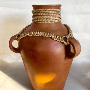 May include: A terracotta-colored vase with two handles and a woven rope detail around the neck. The vase has a rounded body and a slightly flared rim. The rope detail is a light tan color, adding a rustic touch to the pottery.
