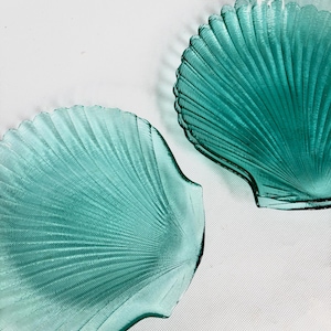 Puede incluir: Dos platos de vidrio en forma de concha, de color turquesa. Los platos tienen una textura acanalada, con las crestas que irradian desde el centro. Los bordes de los platos son festoneados y están sobre un fondo blanco.