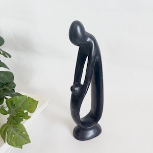 Puede incluir: Una escultura abstracta negra de una persona en una pose curvada, sosteniendo una figura más pequeña. La escultura tiene un acabado liso y pulido y una base redondeada. La figura mide aproximadamente 25 cm de altura.