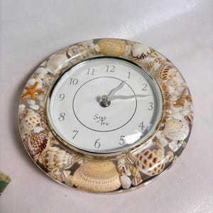 Może przedstawiać: Okrągły zegar ścienny z białą tarczą, czarnymi cyframi i napisem Sands of Time. Zegar jest osadzony w przezroczystej ramie z żywicy, w której zatopiono różnorodne naturalne muszle oraz małą pomarańczową rozgwiazdę w okrągłym układzie.