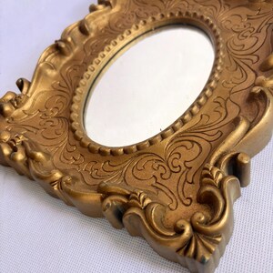 Puede incluir: Un espejo dorado y ornamentado con una superficie ovalada. El marco presenta detalles de volutas y un dise&ntilde;o decorativo texturizado. El espejo est&aacute; en &aacute;ngulo, mostrando los intrincados detalles del marco.