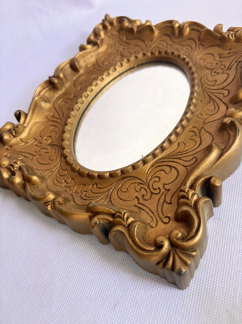Puede incluir: Un espejo dorado y ornamentado con una superficie ovalada. El marco presenta detalles de volutas y un dise&ntilde;o decorativo texturizado. El espejo est&aacute; en &aacute;ngulo, mostrando los intrincados detalles del marco.