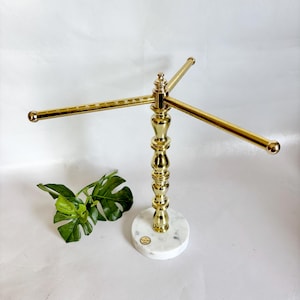 May include: A gold-colored metal jewelry display stand with three horizontal bars. The stand has a decorative, ornate design and sits on a round, white marble base. A green leaf is in the lower left corner.