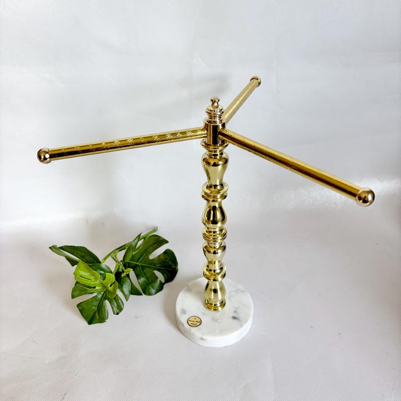 May include: A gold-colored metal jewelry display stand with three horizontal bars. The stand has a decorative, ornate design and sits on a round, white marble base. A green leaf is in the lower left corner.
